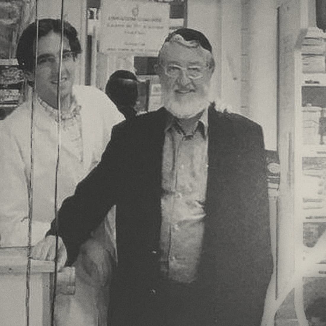 Photographie d'archive en noir et blanc : deux hommes derrière le comptoir d'une charcuterie, l'un en blouse blanche, l'autre en costume, devant les rayonnages de la boutique